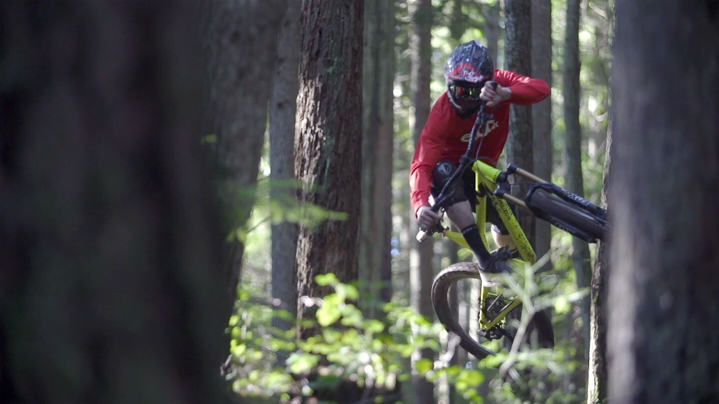 Mountain biker wearing a helmet and red shirt performing a jump in a dense forest, with the bike tilted sideways.